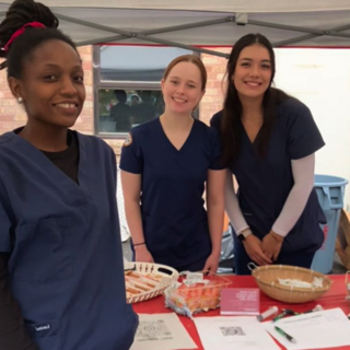 3 nursing club students smile at the camera. They are posing next to a booth in their blue scrubs.